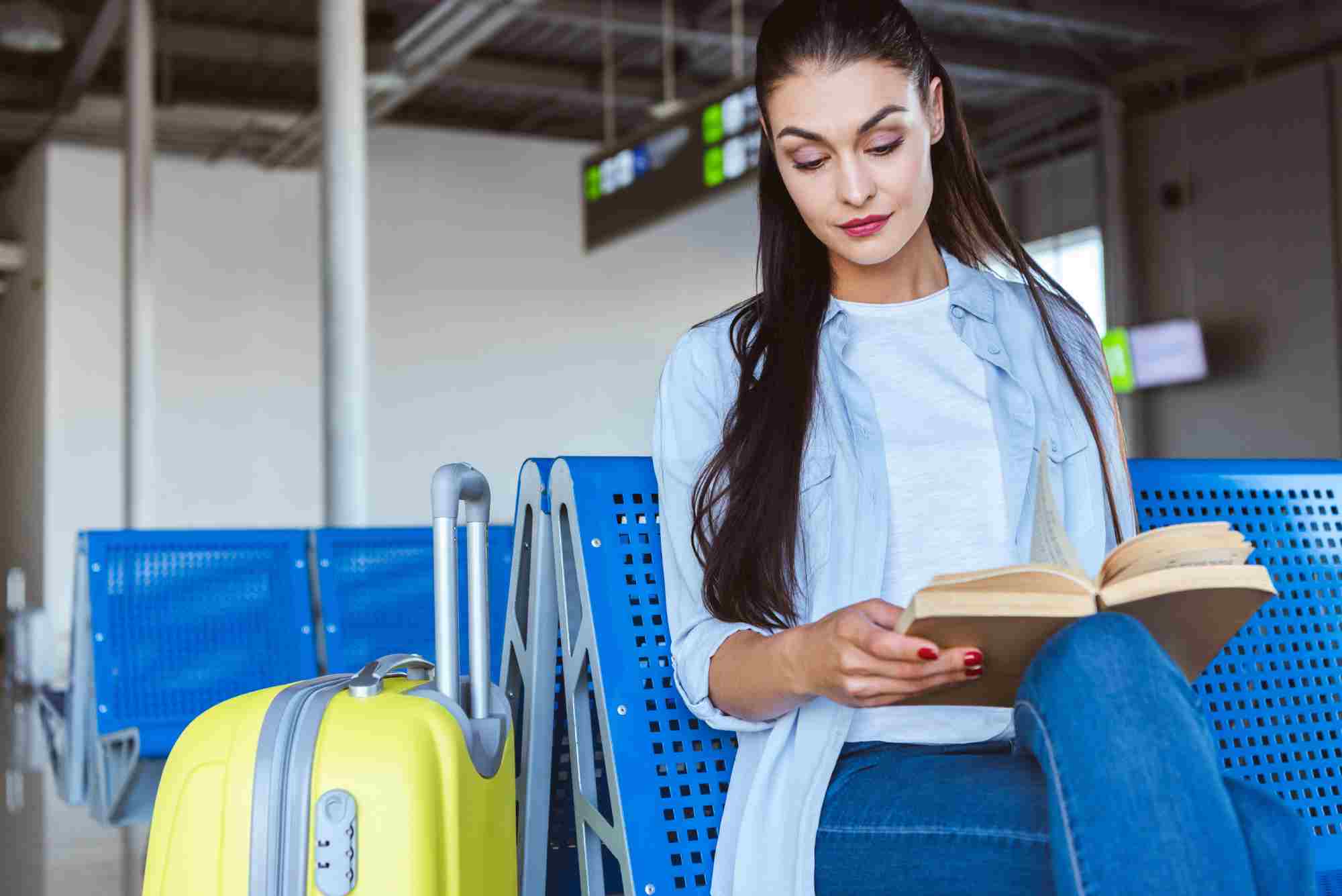 Woman reading a book while sitting at the airport next to her suitcase